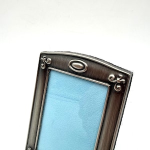 May include: A rectangular picture frame with a brushed dark metallic finish. The frame features decorative scrollwork at the top corners and a centered oval accent. The interior display area is filled with a solid light blue rectangular insert.