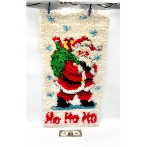 May include: A rectangular latch hook wall hanging featuring a Santa Claus figure in a red suit carrying a green sack of gifts against a cream background with blue snowflakes. The text Ho Ho Ho is in red at the bottom, with a one dollar bill below.