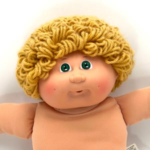 May include: A vintage Cabbage Patch Kid doll with a light skin tone, green eyes, and a knitted yellow yarn hairstyle. The doll has rosy cheeks and a neutral expression. The doll is wearing a peach-colored onesie.