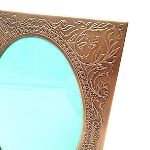 May include: A rectangular picture frame with a metallic bronze finish and an oval center opening. The frame features an intricate embossed pattern of swirling vines, leaves, and small flowers along the corners and edges surrounding the light blue insert.