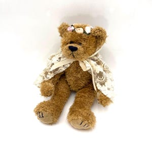 May include: A brown teddy bear with a black nose and a floral crown. The bear is wearing a cream-colored lace shawl tied with a ribbon. The bear is sitting with its legs extended.
