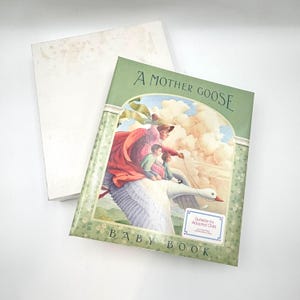 May include: A hardcover baby book titled A Mother Goose Baby Book with a green floral border. The cover illustration depicts a person in a red cloak riding a large white goose with two children. A small white label reads Suitable for Adopted Child.