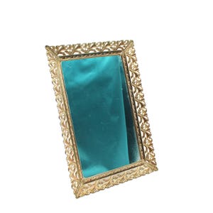 May include: A rectangular gold-colored mirror with an ornate, floral-patterned frame. The mirror's surface is a deep teal color. The frame has a detailed, vintage design, adding to its decorative appeal.