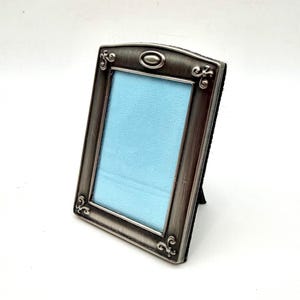 May include: A rectangular picture frame with a brushed dark metallic finish. The frame features an arched top with a central oval accent and decorative scrollwork at the corners. A light blue insert is visible within the frame opening.