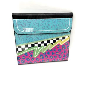 May include: A Trapper Keeper binder featuring a teal top section with purple geometric shapes, a black and white checkered middle band, and a bottom section with a pink and blue leopard print pattern. A bright green zigzag line crosses the front.