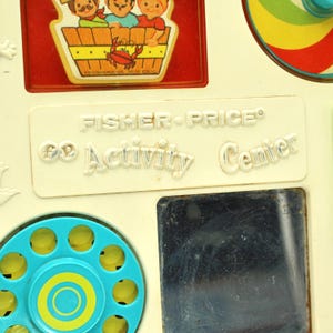 May include: A vintage Fisher-Price "Activity Center" toy with a blue and yellow rotary phone, a mirror, and a spinning top. The toy is made of white plastic and has a red and white border around the edges.