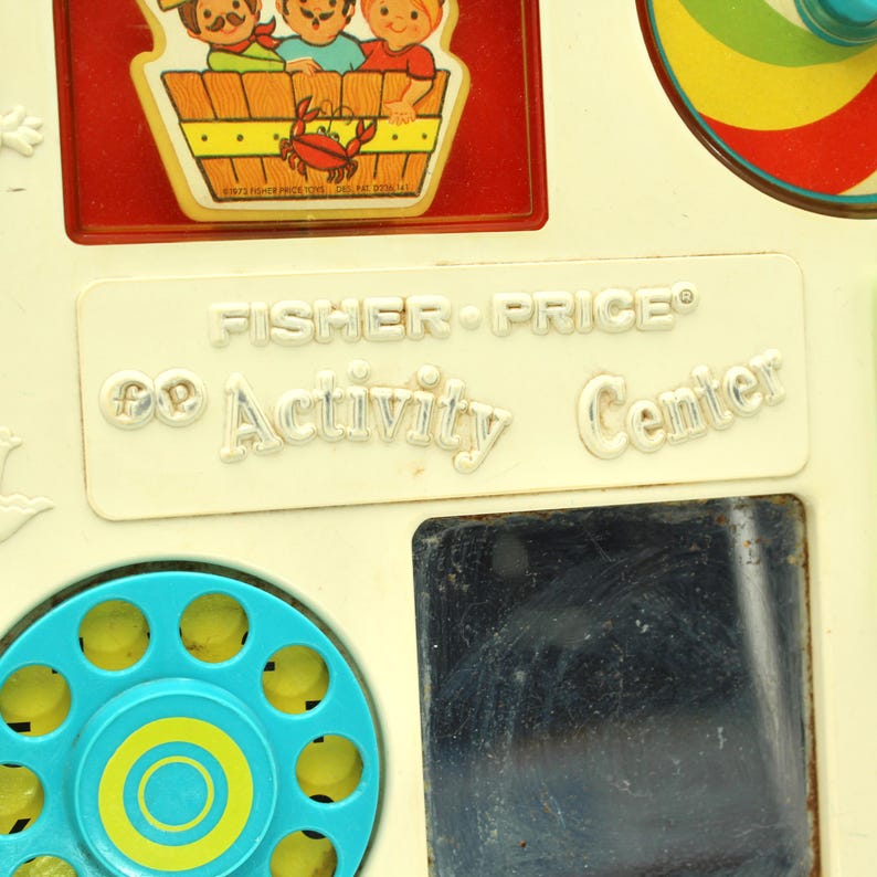 May include: A vintage Fisher-Price "Activity Center" toy with a blue and yellow rotary phone, a mirror, and a spinning top. The toy is made of white plastic and has a red and white border around the edges.
