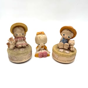 May include: Three ceramic figurines featuring two children sitting on circular bases with a squirrel and a cat, and one smaller child sitting between them. The figures have blonde hair, pastel-colored clothing, and painted facial features.