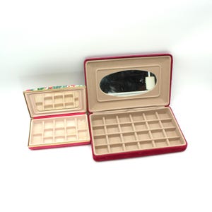 May include: Two open jewelry boxes. The larger box is red with a beige interior and a mirror in the lid. The smaller box has a floral pattern on the lid and a pink interior. Both boxes have compartments for jewelry.