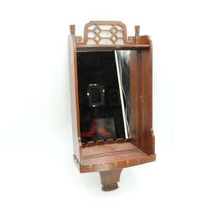 May include: A vintage wooden wall mirror with a shelf and decorative fretwork. The mirror is rectangular and framed in dark brown wood. The shelf is curved and has a decorative edge. The mirror has a decorative base.