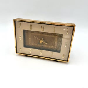 May include: A vintage gold-toned rectangular clock with a light brown face and gold numerals. The clock has a rectangular patterned center and gold hands. The clock is a classic design, likely from the mid-20th century.