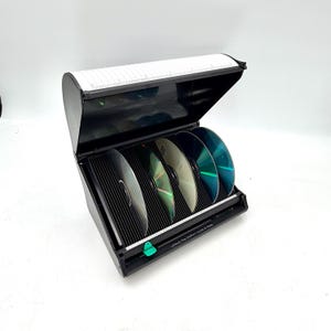 May include: Black CD storage case with a hinged lid, holding several CDs. The case has a ribbed interior and a white label on the lid with numbers. The text "Unlock the bottom locks & Press" is visible. A green button is on the front.
