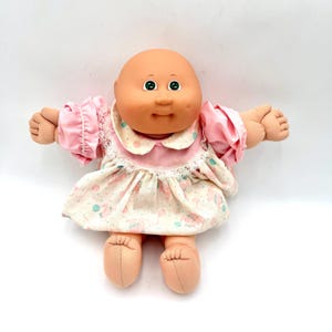 May include: A bald doll with a round peach-toned head, green eyes, and dimpled cheeks. The doll wears a cream-colored dress featuring a pattern of small pink and teal balloons, accented with a pink collar and white lace trim on the sleeves.