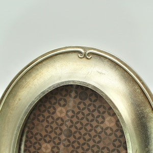May include: Oval silver-toned picture frame with a decorative top detail. The frame has a slightly distressed finish and a patterned background visible through the opening. A classic design for displaying photographs or artwork.