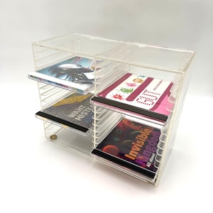 May include: A clear acrylic CD storage rack with two vertical columns and multiple horizontal slots. The rack holds four CD cases, including titles such as Invisible Kingdom, and features a transparent, modular design for organizing and displaying media.