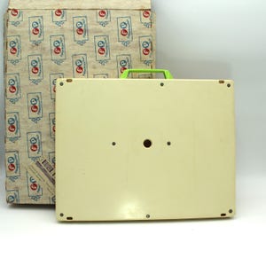 May include: The bottom of a white plastic toy with a green handle. The toy has a round hole in the center and four small holes around the edges.
