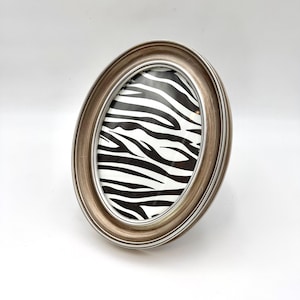 May include: An oval-shaped picture frame with a metallic, brushed bronze finish and a raised, tiered border. The frame contains a decorative insert featuring a high-contrast black and white zebra stripe pattern that fills the entire oval display area.