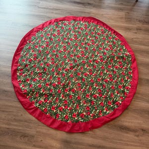 May include: A circular Christmas tree skirt featuring a festive pattern of red apples, green holly leaves, and brown pinecones on a cream background. The skirt has a solid red fabric border around the edge and a straight slit for placement.