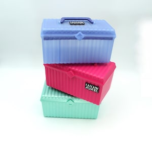 May include: Three stackable plastic storage boxes with a ribbed design. The top box is light blue, the middle box is pink, and the bottom box is light green. Each box has a black label with the word "SASSY" printed on it.
