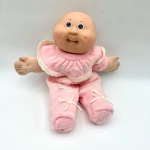 May include: A soft-sculpture doll with a round, peach-toned face, dimpled cheeks, and dark eyes. The doll wears a light pink knitted outfit featuring a ruffled collar with white trim, embroidered floral details, and small white ribbon bows on the legs.