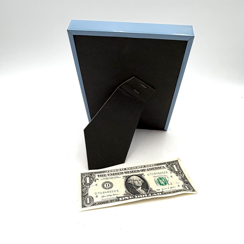 May include: A pale blue picture frame with a black backing and a black easel stand. A one-dollar bill is placed in front of the frame. The frame is rectangular and has a simple, modern design.