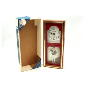 May include: A rectangular wooden wall clock with a red face and a photo frame. The clock face has black numbers and hands. The photo frame displays a picture of a person. The clock is in its original box, which is blue and white.