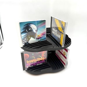 May include: A black plastic rotating CD storage rack holding four compact discs. The discs feature cover art including a robotic figure, a yellow textured design with the text WAHT MISTEAK?, a colorful graphic with the text Invisible Kingdom, and a white cover with the text GUT.