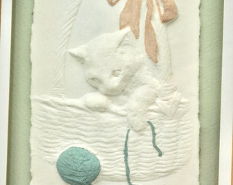 May include: A white kitten sits in a woven basket with a light blue ball of yarn and a pink bow. The kitten is looking down at the yarn. The image is in a white frame with a green mat.