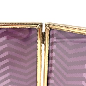 May include: A gold-colored, hinged picture frame with a decorative rope-like border. The frame is open, revealing a purple and gray chevron pattern. The frame is designed to hold two photos.