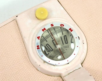May include: A vintage pink Hanson bathroom scale with a round dial face. The dial face has a black needle and white numbers. The scale reads "HANSON NOT LEGAL FOR TRADE MADE IN U.S.A. PAT. PENDING".