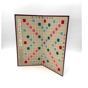 May include: A Scrabble board game with a tan playing surface, featuring a grid of squares with pink, blue, and red bonus spaces. The board is open, displaying the game's name and letter values. The border is a dark burgundy color.