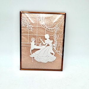 May include: A framed silhouette art piece featuring a white paper cut-out of a woman seated at a table, reading. The background is a textured, peach-colored material with a window and curtain design. The frame is a dark brown color.