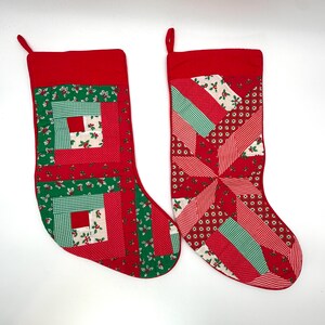 May include: Two Christmas stockings featuring patchwork designs in red, green, and white. The left stocking has a square block pattern, while the right stocking displays a star-like geometric arrangement. Both have solid red cuffs and hanging loops.