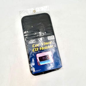 May include: A black car visor CD holder in a clear plastic package. The holder is rectangular and features the text "Car Visor CD Holder" and holds 10 CDs. The package has a small hole at the top for hanging.