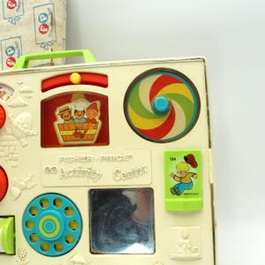 May include: Vintage Fisher-Price 60 Activity Center toy with a white plastic base, featuring a spinning wheel, a mirror, a phone dial, and a sliding panel with a picture of three children. The toy also has a red and yellow slider, a green and yellow spinning wheel, and a green card with a boy on it.