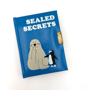 May include: A blue hardcover journal with the text SEALED SECRETS printed in white. The cover features a light brown seal holding a flipper to its mouth and a small black and white penguin. A metallic gold-colored latch is attached to the side.