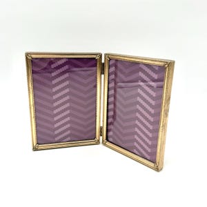 May include: A hinged, gold-toned metal picture frame, open to display two purple fabric panels with a chevron pattern. The frame has a decorative rope-like border and is set against a white background.