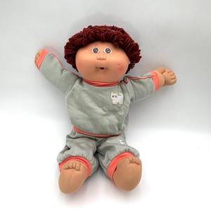 May include: A doll with a round face, rosy cheeks, and dark reddish-brown yarn hair. The doll wears a light gray long-sleeved top and matching pants with coral-colored trim at the cuffs and waist. A small white cat graphic is on the chest.