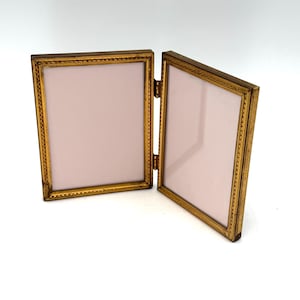 May include: A hinged, gold-colored picture frame with two rectangular openings. The frame has a decorative border and a light pink background. The frame is open, revealing the interior. The hinges are visible.