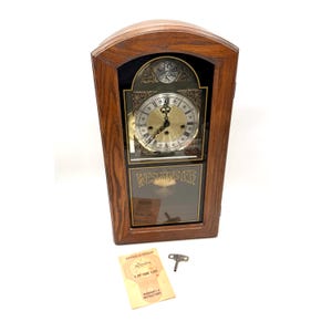 May include: A wooden Westminster clock with a gold-colored clock face and black hands. The clock has a glass front and a dark brown wooden frame. The clock face has Roman numerals. A key and a warranty card are next to the clock.