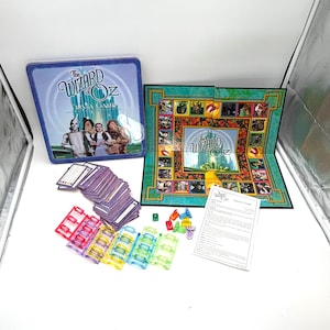 May include: A Wizard of Oz trivia game set. The game includes a blue tin with the game title and characters, a game board, cards, dice, and game pieces. The board is green with images from the movie.