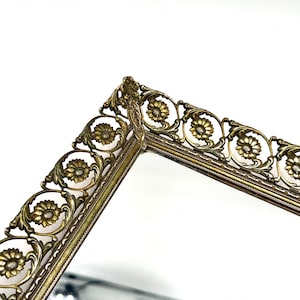 May include: Close-up of a vintage vanity mirror with a decorative gold-toned metal frame. The frame features a floral design with detailed embellishments. The mirror's surface is reflective, and the overall aesthetic is ornate and elegant.