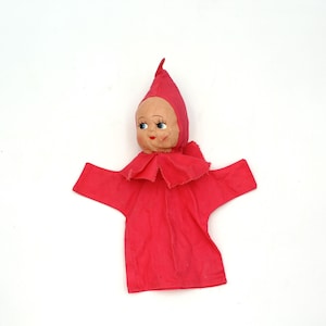 May include: A vintage hand puppet featuring a painted doll face with blue eyes and a red pointed hat. The puppet has a red fabric body with a ruffled collar and outstretched arms, set against a white background.