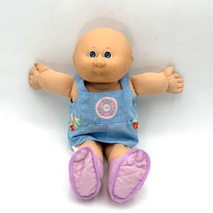 May include: A bald doll with a round head, blue eyes, and a peach-toned complexion. The doll wears light blue denim-style overalls featuring a repeating CPK logo pattern, a circular pink and white chest patch, and quilted light purple shoes.