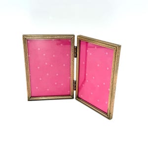 May include: A hinged, gold-colored metal picture frame with two rectangular openings. Each opening features a pink background with white polka dots. The frame is open, revealing the interior design. The frame is on a white background.