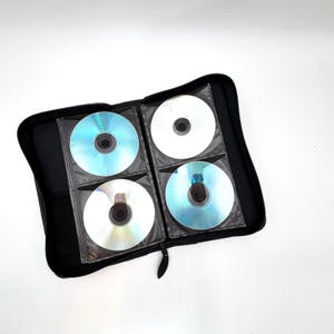 May include: A black zippered storage case open to reveal four circular discs held in individual clear plastic sleeves. The discs have reflective surfaces with iridescent blue and silver tones, arranged in a two-by-two grid inside the case.
