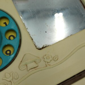 May include: A vintage toy telephone with a blue rotary dial and a small mirror. The phone has a cream-colored base with a mushroom and flower design.