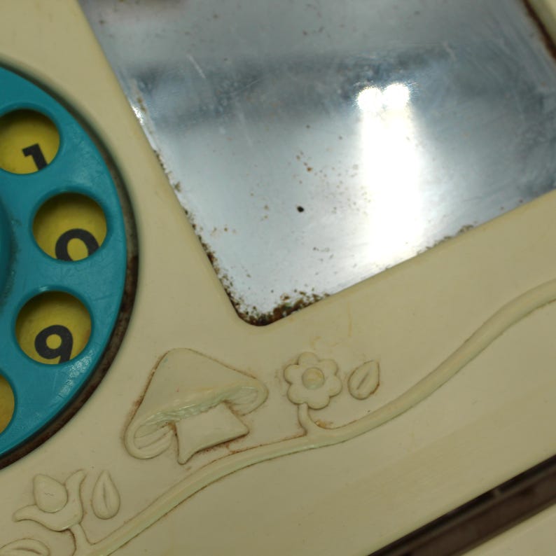 May include: A vintage toy telephone with a blue rotary dial and a small mirror. The phone has a cream-colored base with a mushroom and flower design.