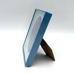 May include: A blue picture frame with a floral design. The frame is angled, showing a wooden easel back. The artwork features a pattern of red, yellow, and blue flowers on a light blue background.