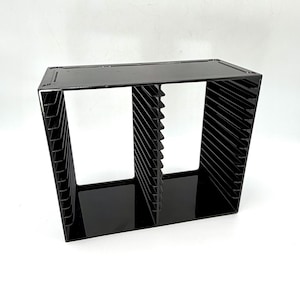 May include: A black acrylic storage rack featuring two vertical sections with multiple horizontal slots for organizing items. The unit has a solid top and base with a glossy finish, designed with evenly spaced dividers to hold flat objects securely.